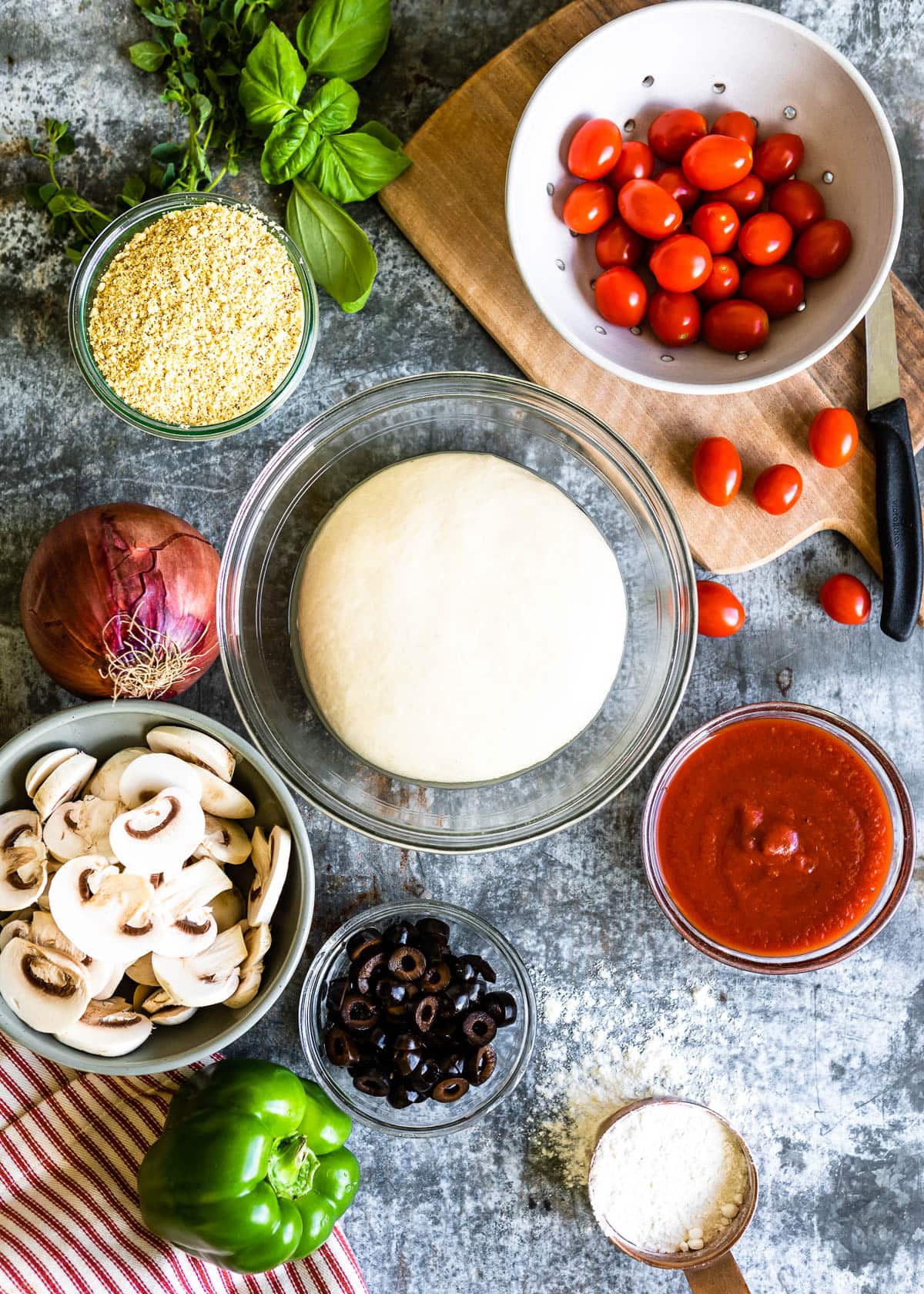 several ingredients in bowls like fresh tomatoes, vegan parmesan cheese, purple onion, black olives, marinara sauce, and gluten-free dough.