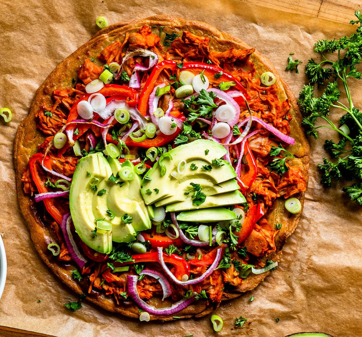 jackfruit BBQ pizza on parchment, topped with fresh avocado, purple onions and red peppers.