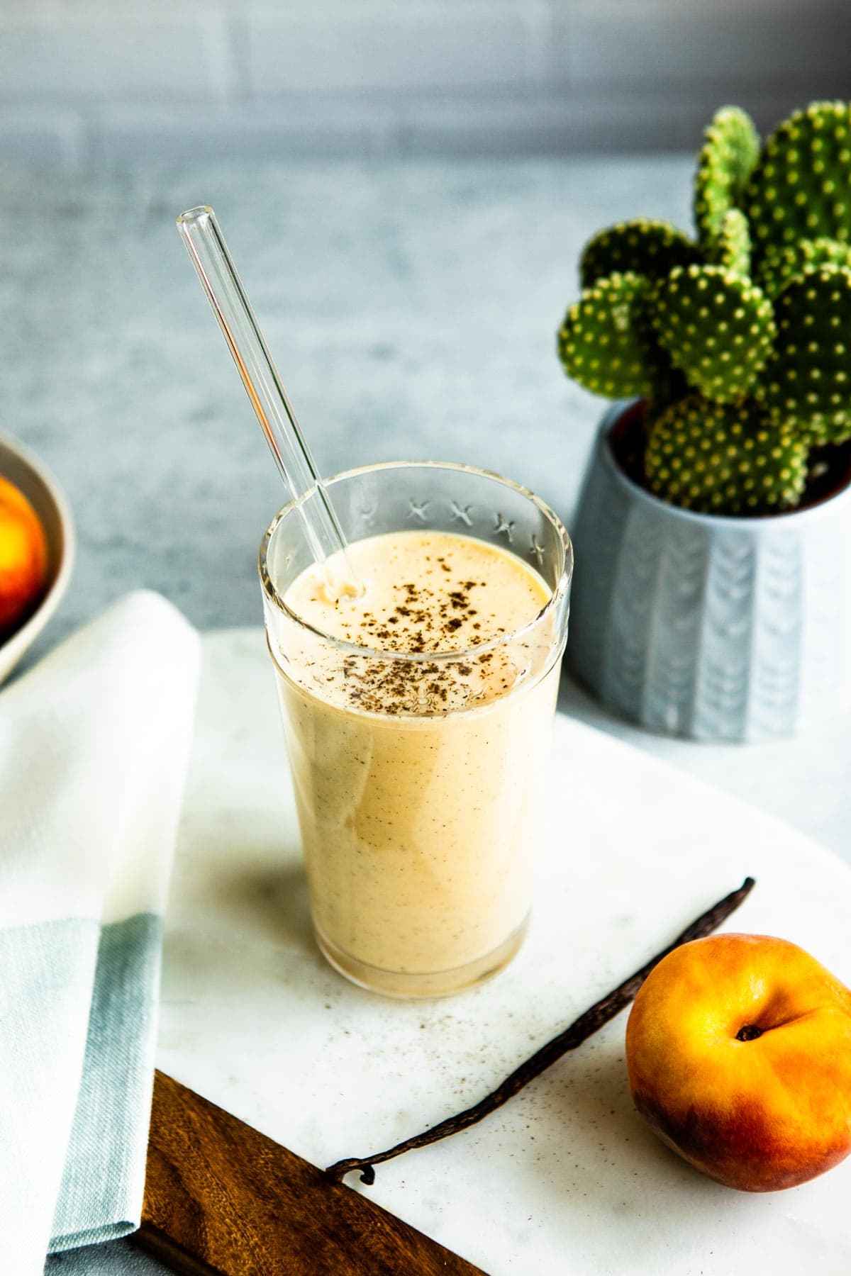 healthy protein shakes in a glass with a glass straw on a marble slab with a vanilla bean and whole peach sitting next to it.
