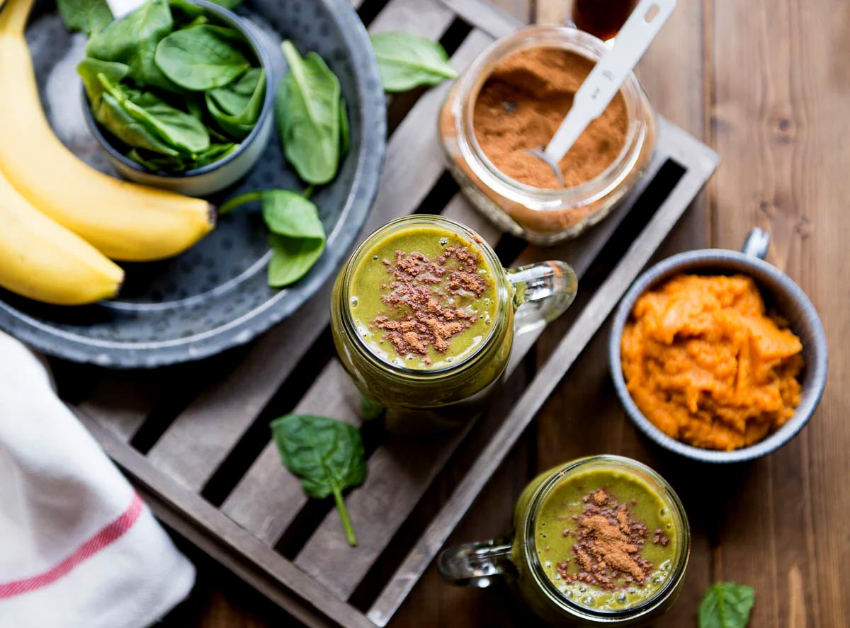 Two glass mugs of pumpkin smoothie next to a mug of pumpkin puree and a jar of spice, with blurred bananas and spinach on a plate.