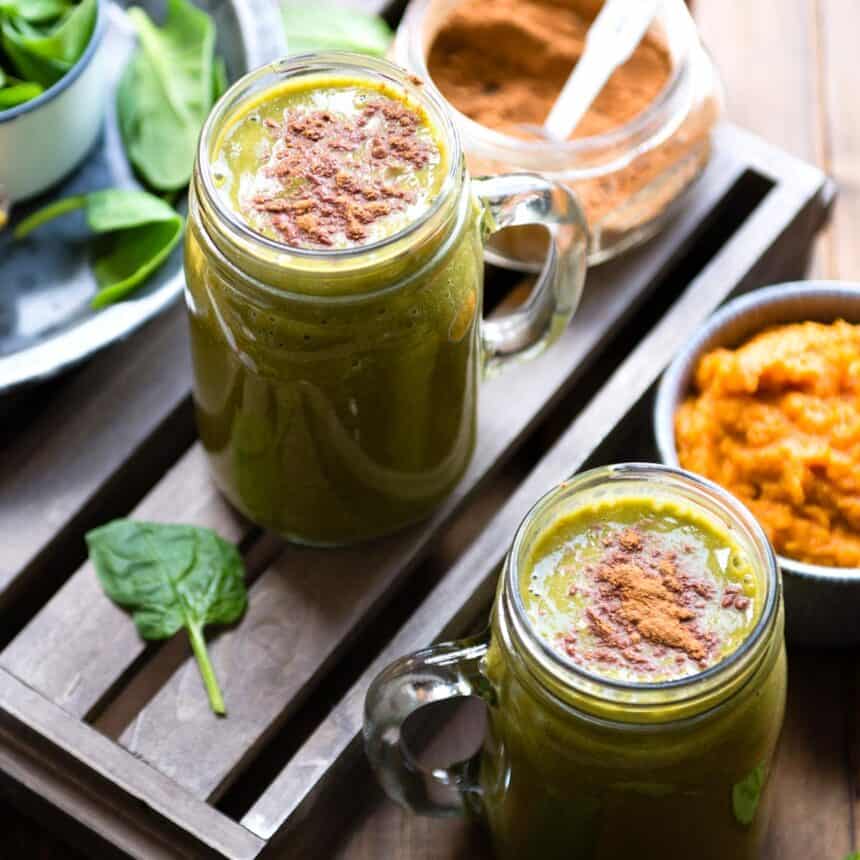 Glass mugs with pumpkin smoothie topped with pumpkin pie spice next to a mug of pumpkin puree and a jar of spice.