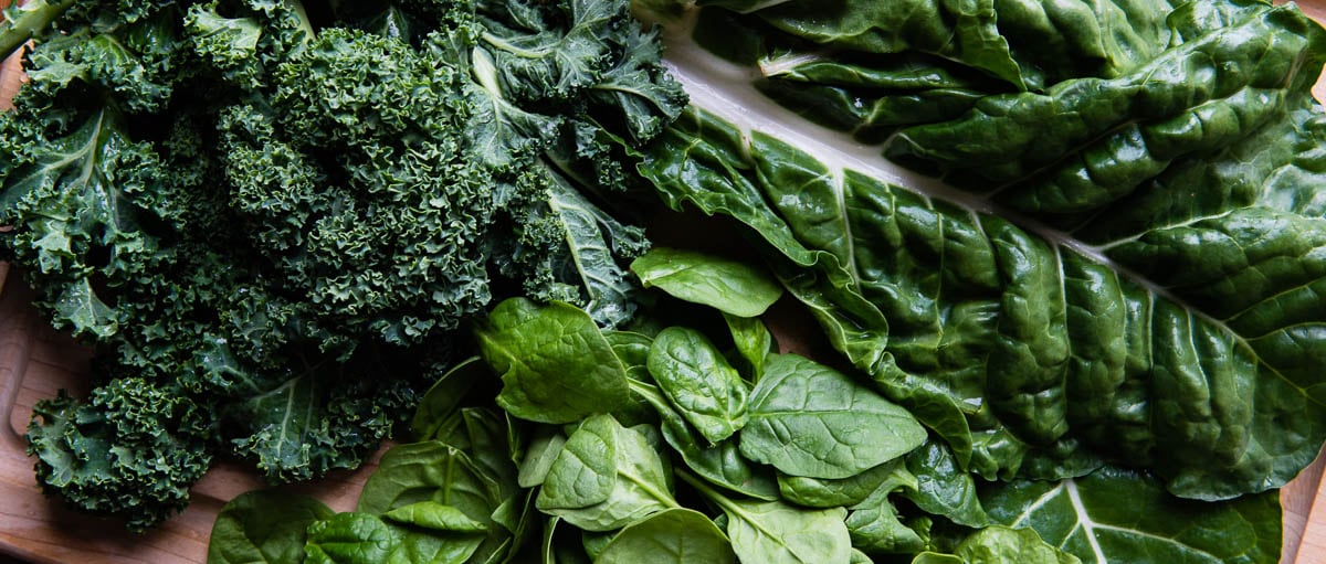 Different types of leafy greens including kale, Swiss chard and spinach, laying on top of each other.