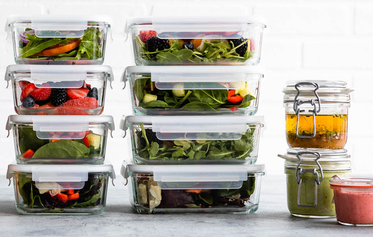8 stacked glass containers of salad next to 3 round glass containers of dressing showing how to use leftovers as healthy snacks for work.