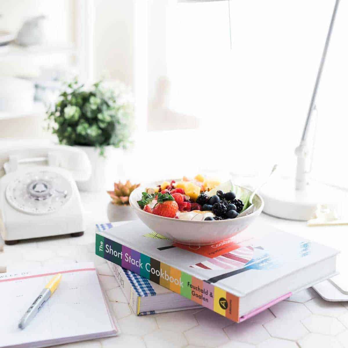 white bowl of fruits and nuts on top of 2 books on a white desk.