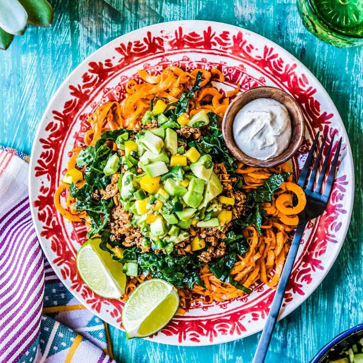 sweet potato noodles topped with kale, avocado, mango on a red and white plate.