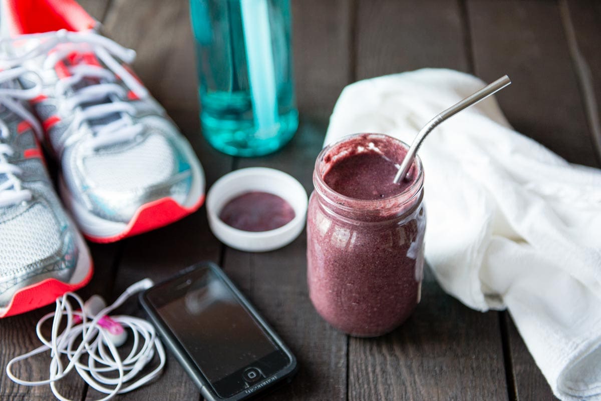 purple smoothie in mason jar with stainless straw next to a phone, headphones and running shoes.