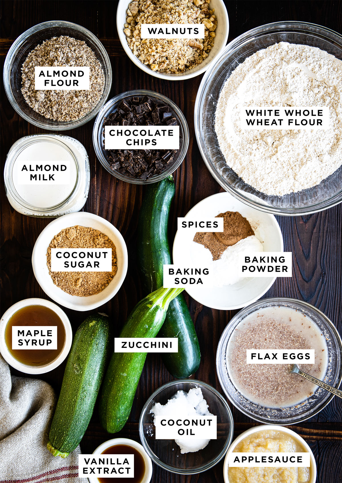 labeled ingredients for zucchini bread recipe including walnuts, almond flour, chocolate chips, white whole wheat flour, almond milk, coconut sugar, spices, baking soda, baking powder, maple syrup, zucchini, flax eggs, coconut oil, vanilla extract and applesauce.