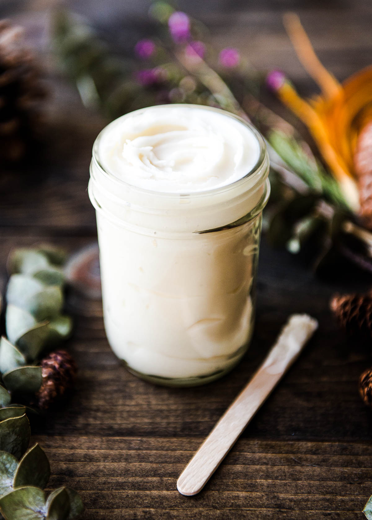 Glass jar of homemade deodorant made with coconut oil and essential oils, with a wooden popsicle stick applicator beside it. A colorful, woodsy arrangement is blurred in the background.