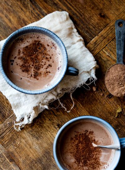 A blue mug filled with rich homemade hot chocolate sits on the countertop, accompanied by a tablespoon of cacao powder and vegan milk.