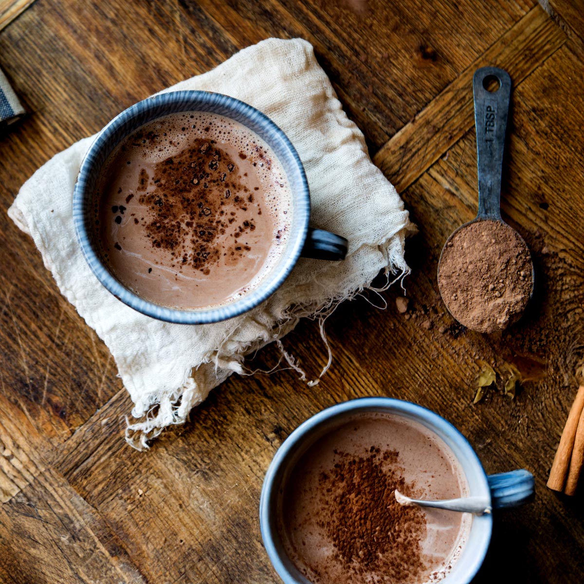 A blue mug filled with rich homemade hot chocolate sits on the countertop, accompanied by a tablespoon of cacao powder and vegan milk.