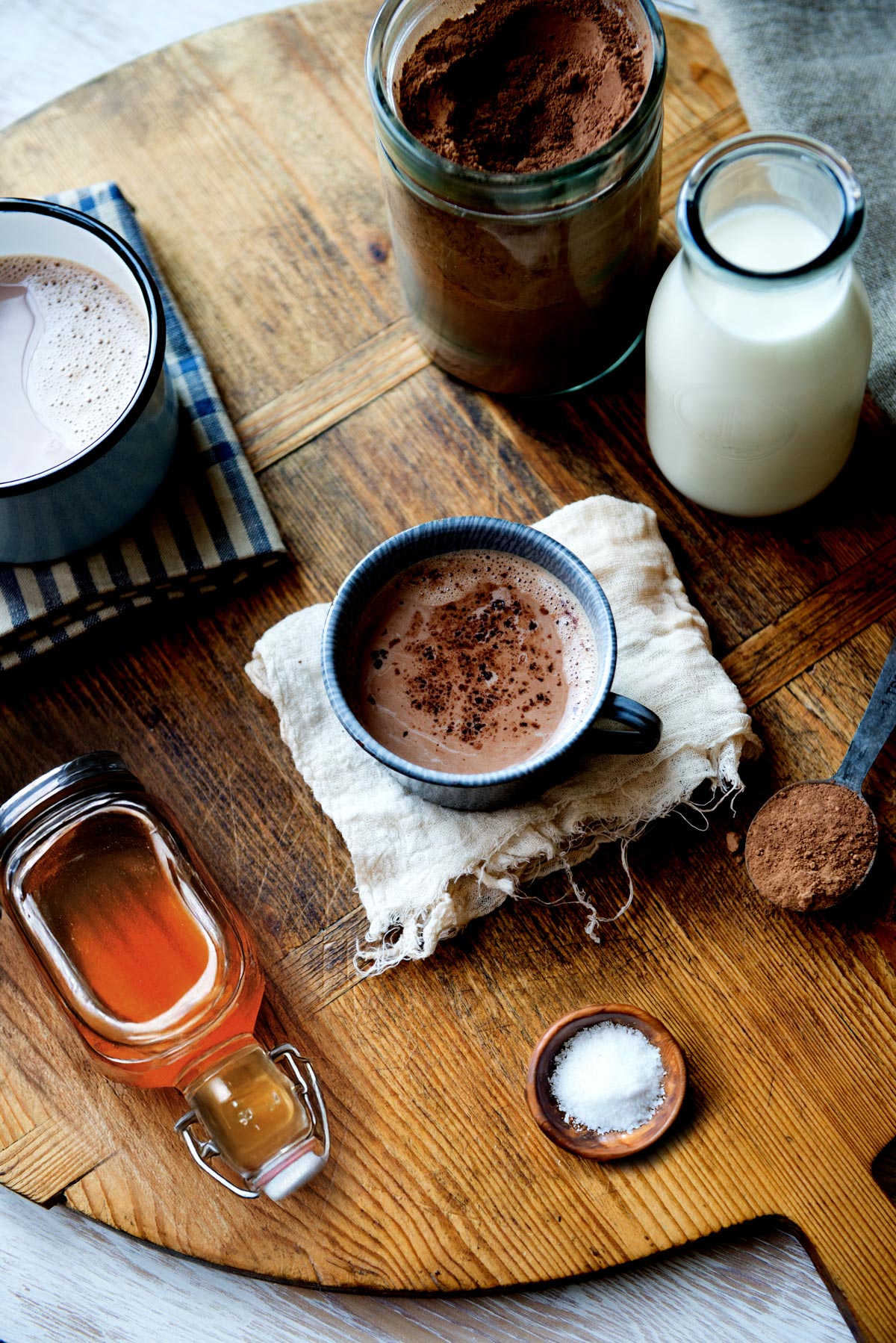 A blue mug holds a vegan hot chocolate recipe, resting on a circular wooden board surrounded by essential healthy hot chocolate ingredients.