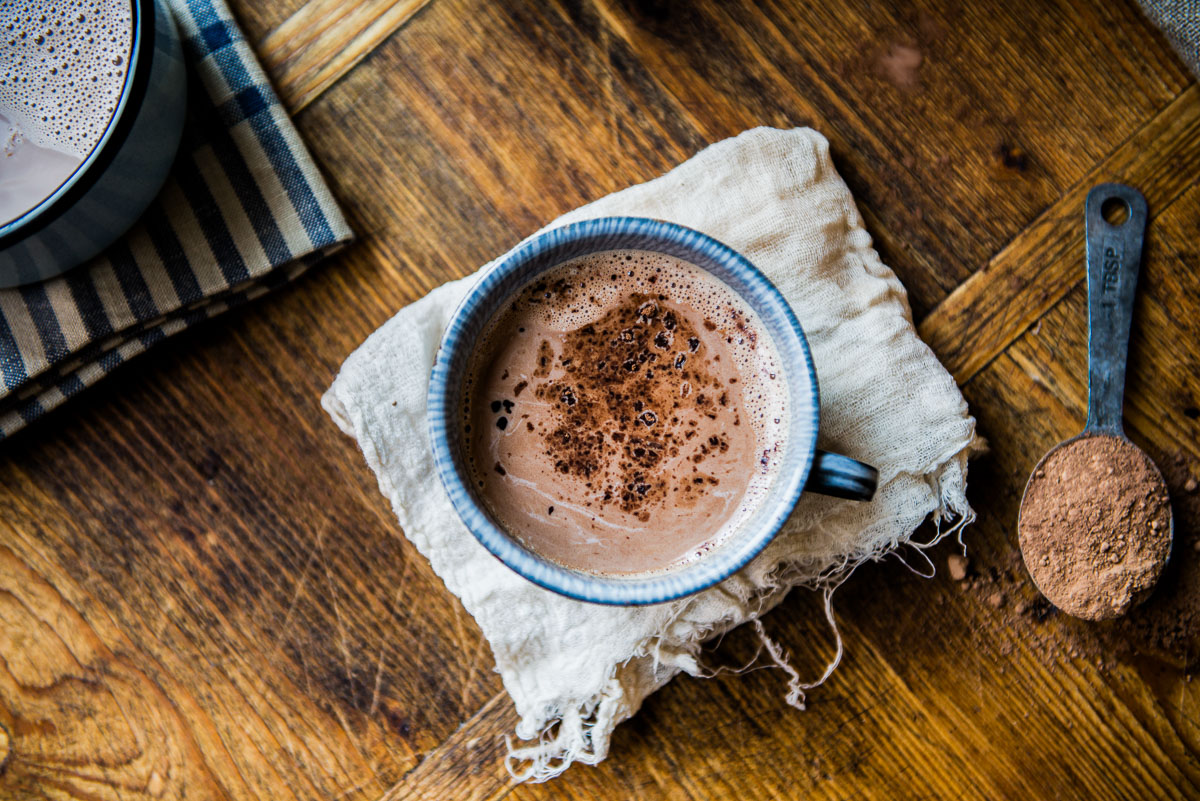 A mug of dairy-free hot chocolate rests on a cheesecloth, next to a tablespoon of cacao powder.