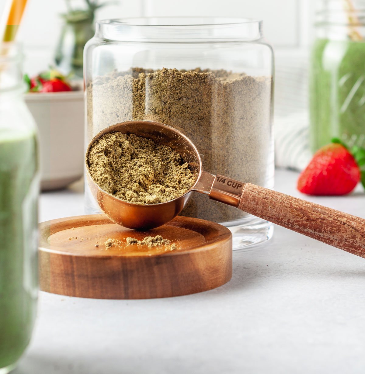 Scoop of homemade protein powder in a 1/4 cup measuring spoon in front of a large glass jar of the full batch of DIY protein powder.