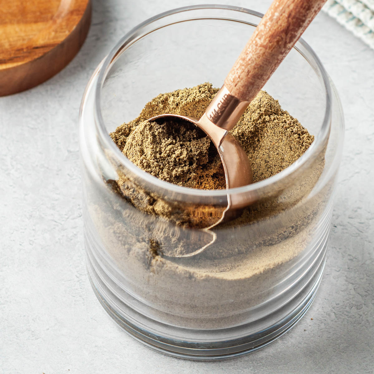 Glass container of DIY protein powder on a grey countertop with a copper measuring spoon scooping out a full scoop. 