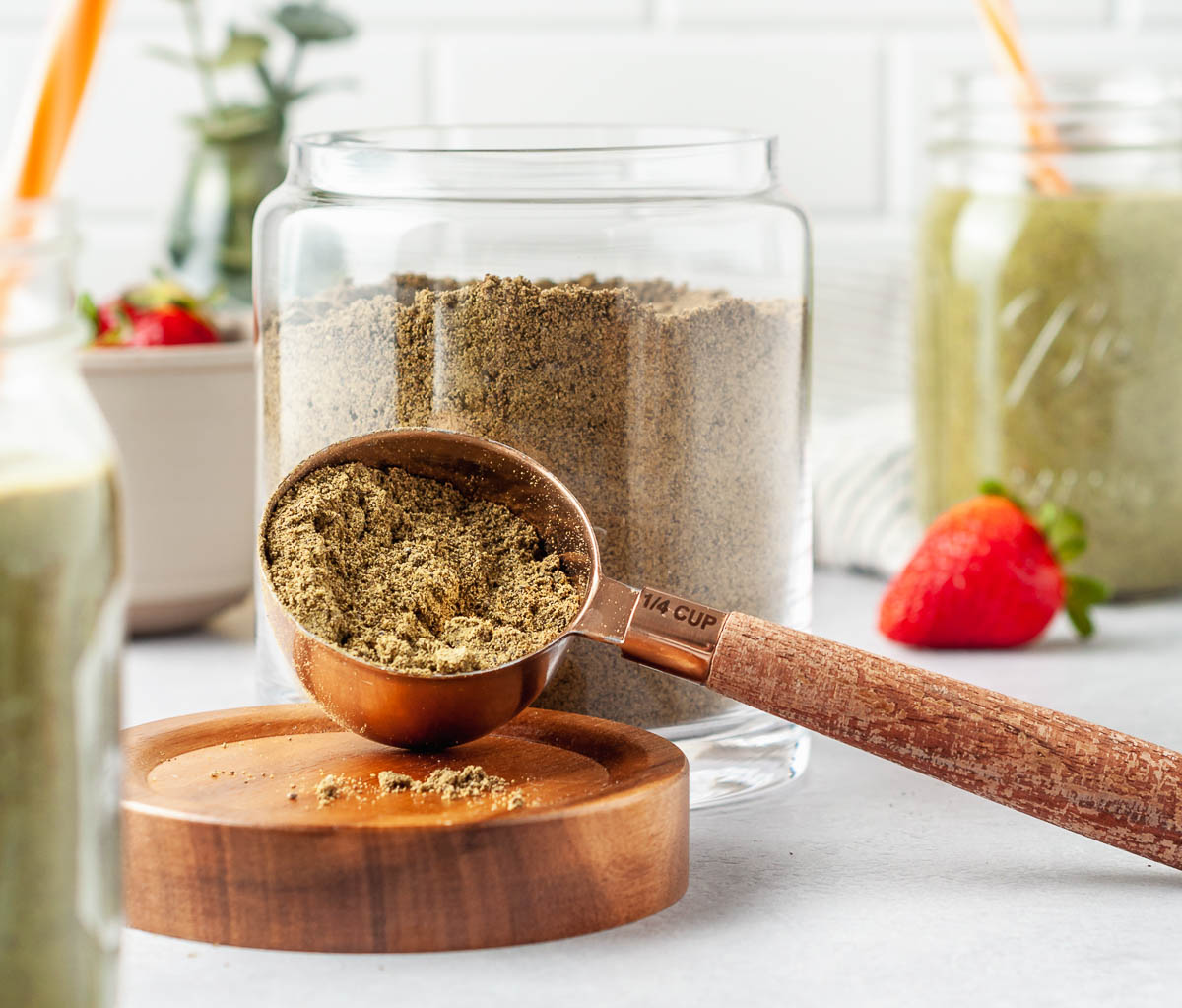 Copper and wooden scoop full of diy protein powder recipe, next to an open glass jar of protein powder surrounded by smoothies and strawberries.