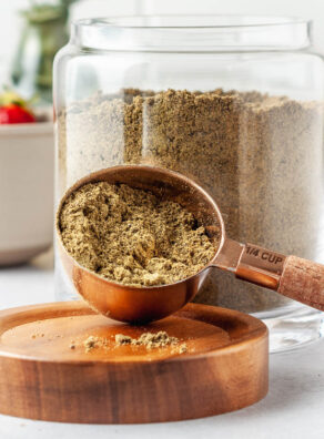 Scoop of homemade protein powder in a 1/4 cup measuring spoon in front of a large glass jar of the full batch of DIY protein powder.