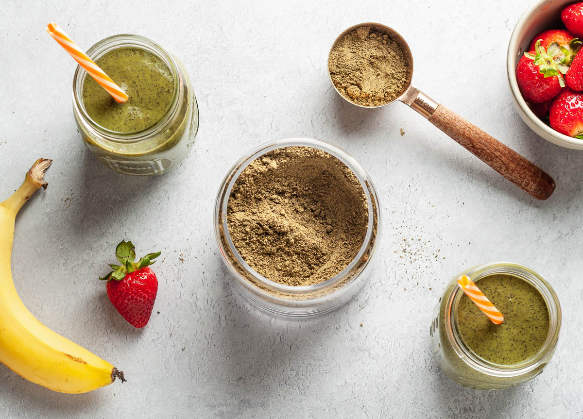 homemade protein powder in between 2 glass jars of green smoothie, both with orange straws.