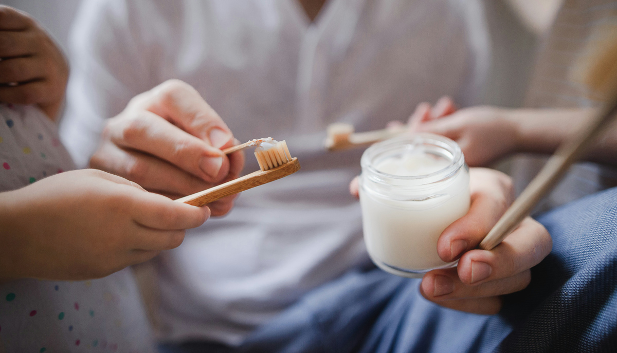 Hands holding toothbrushes around a glass jar filled with homemade toothpaste.