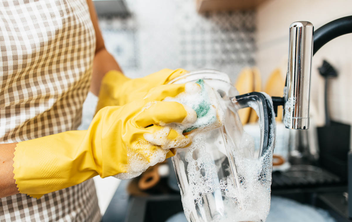 yellow cleaning gloves scrubbing a blender container with a sponge and soap in a sink.