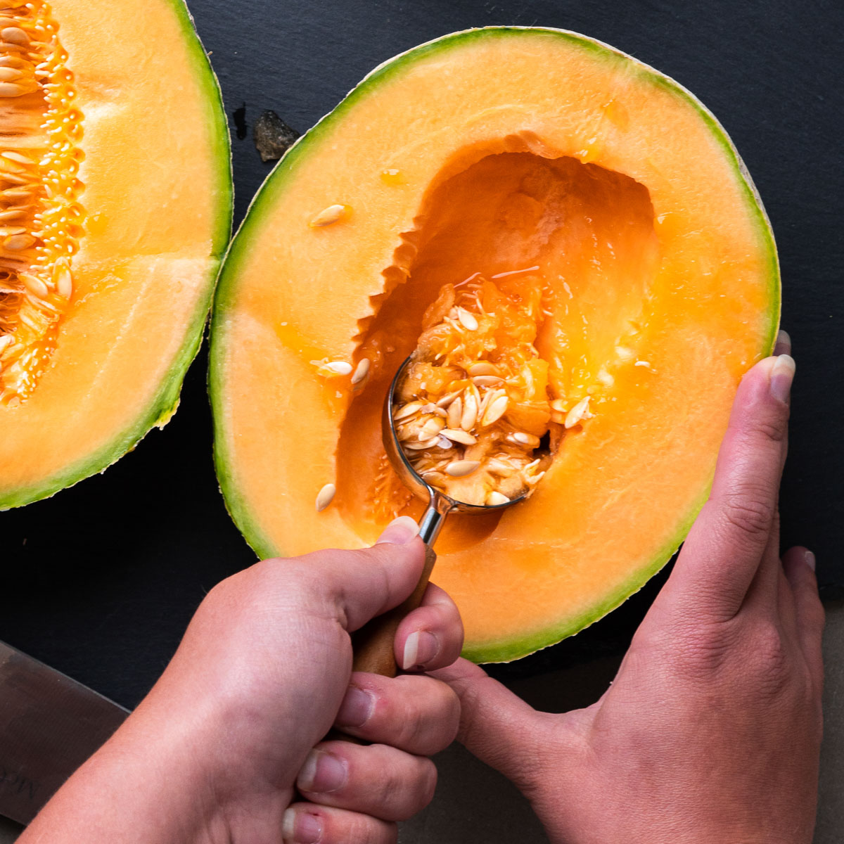 A hand securely holding a cantaloupe as another hand, with a spoon, scrapes out the seeds.