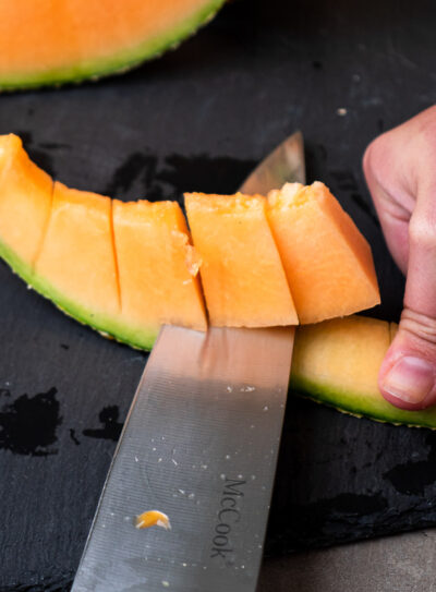 A hand secures the cantaloupe rind while a knife cuts through the melon.