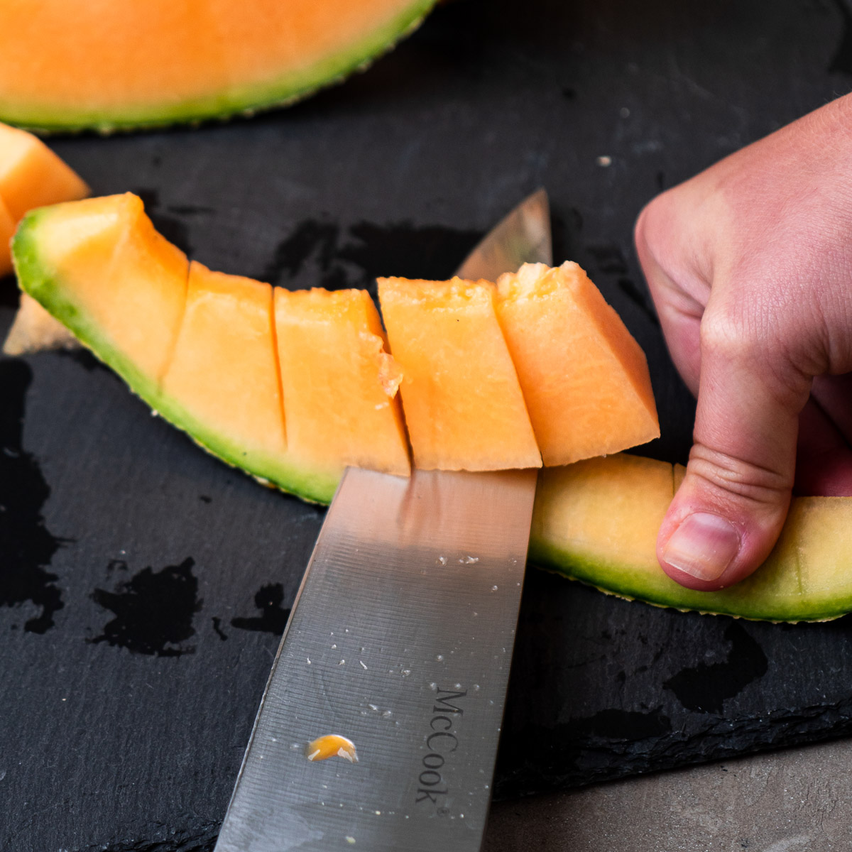 A hand secures the cantaloupe rind while a knife cuts through the melon.
