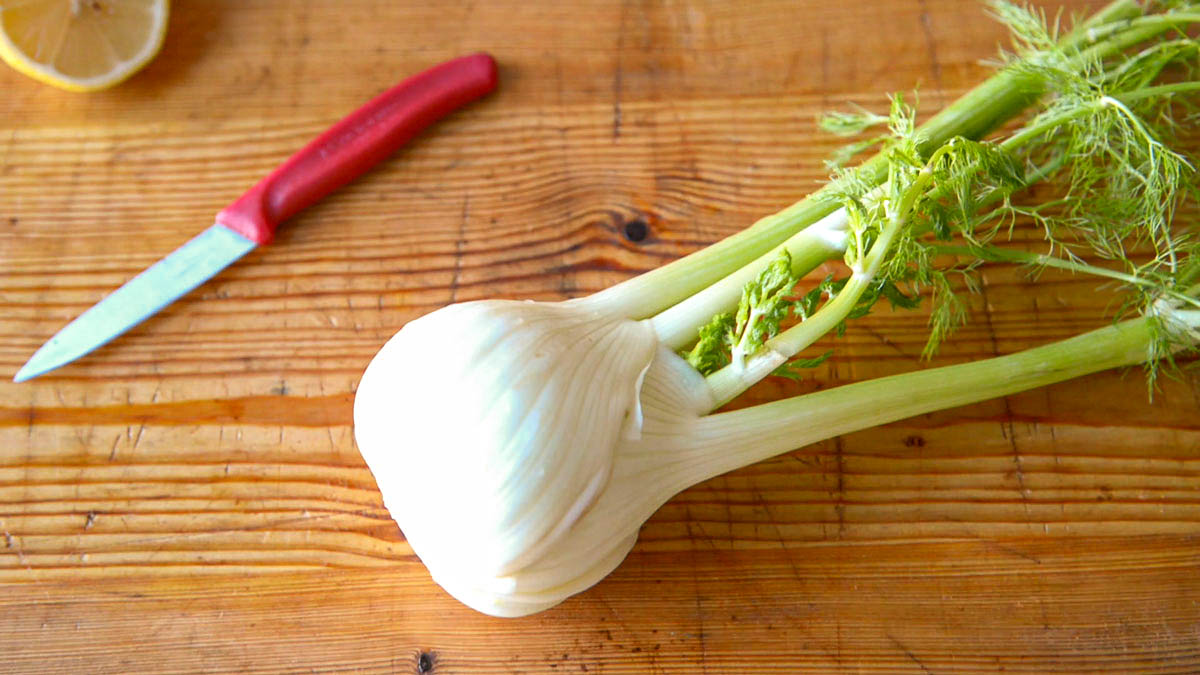 fresh fennel on a wooden cutting board next to a paring knife with a red handle.