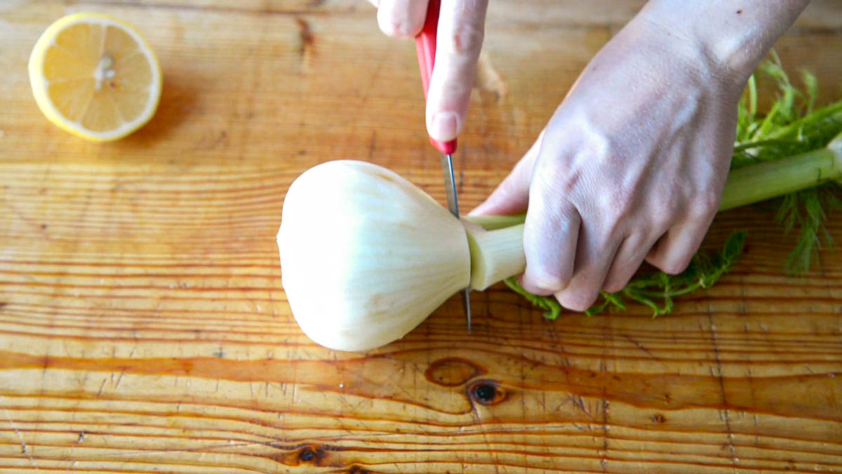 2 hands showing how to cut fennel by first removing the leafy fronds from the white base using a paring knife, next to a cut lemon.