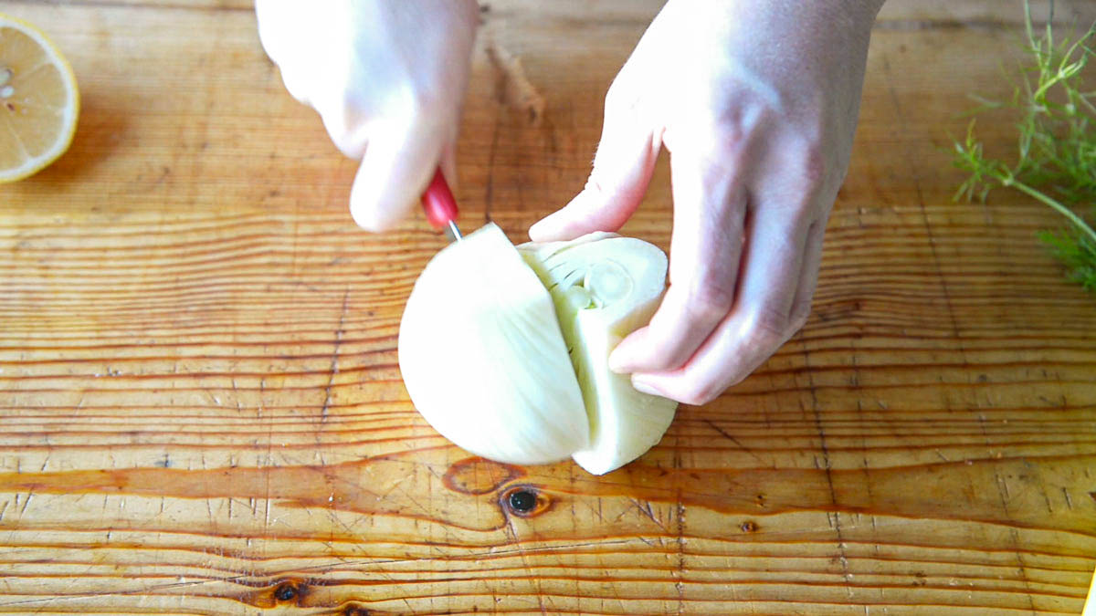 2 hands cutting the white bulb of a fennel in half with a red handled paring knife on a wooden cutting board.