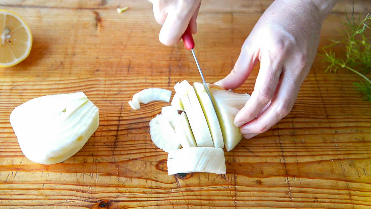 2 hands dicing a fennel bulb on a wooden cutting board using a paring knife with a red handle.