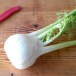 fresh fennel on a wooden cutting board next to a paring knife with a red handle.