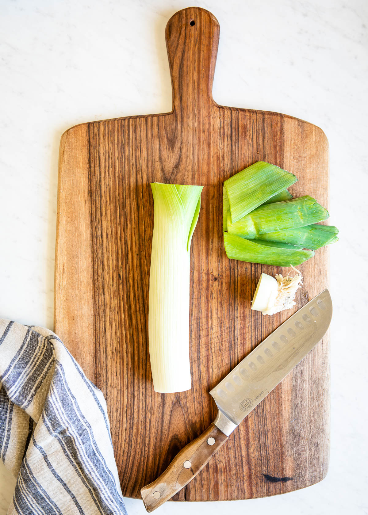 step one of how to cut leeks showing the green top chopped off the white neck on a wooden cutting board with a chef's knife.