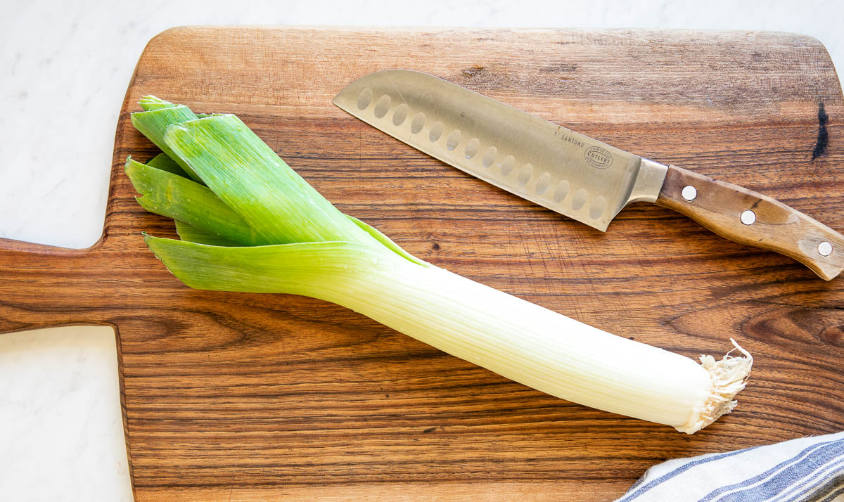 whole leek on a wooden cutting board next to a chef's knife.
