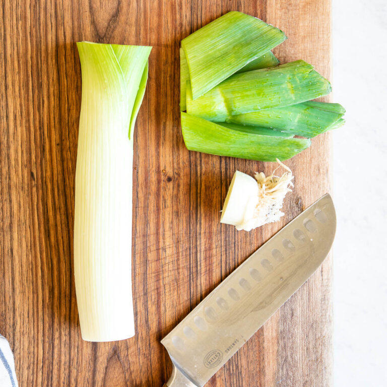 partially cut leek on a wooden cutting board next to a chef's knife.