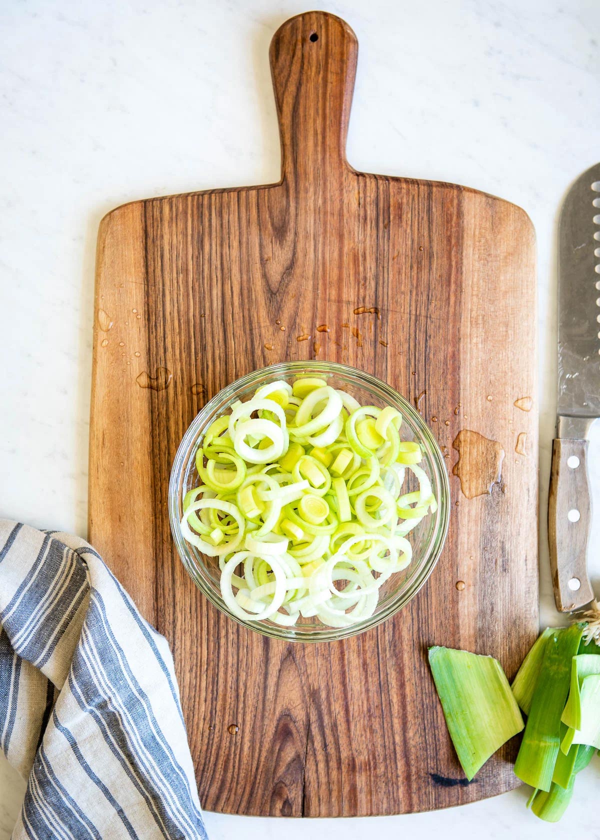 showing how to clean leeks by placing the sliced white circles in a glass bowl full of water, sitting on a wooden cutting board.