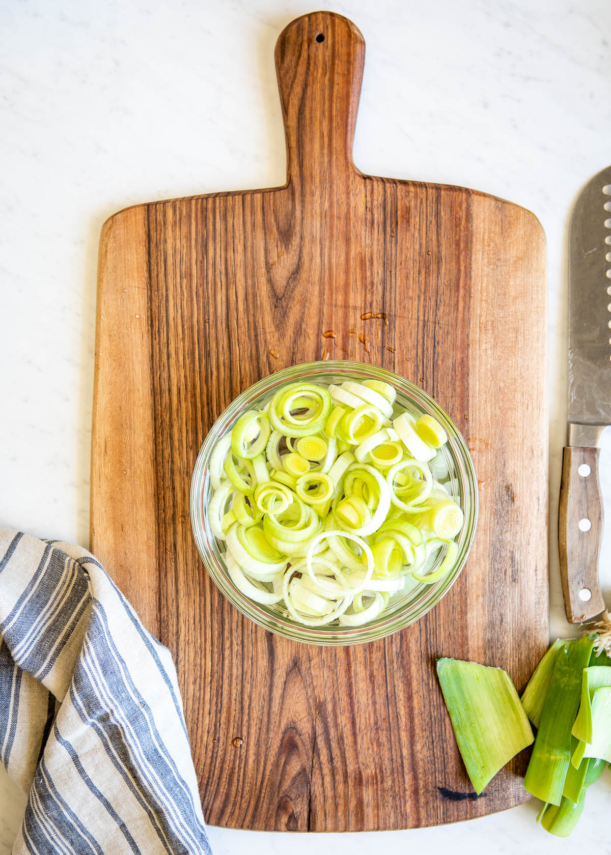 showing how to clean leeks by placing the sliced white circles in a glass bowl full of water, sitting on a wooden cutting board.