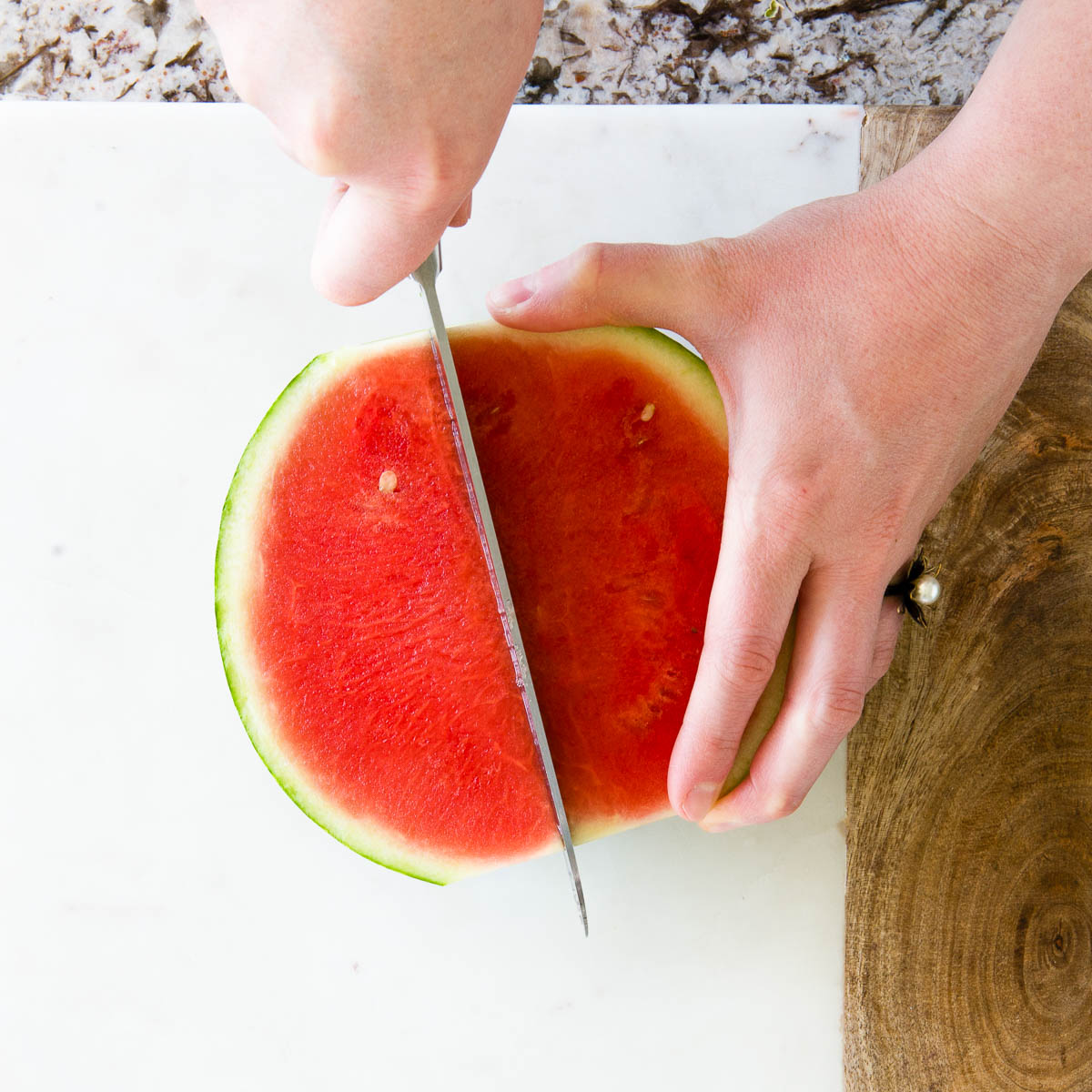 showing how to cut a watermelon by slicing 1/2 into 2 pieces on a marble and wood cutting board.