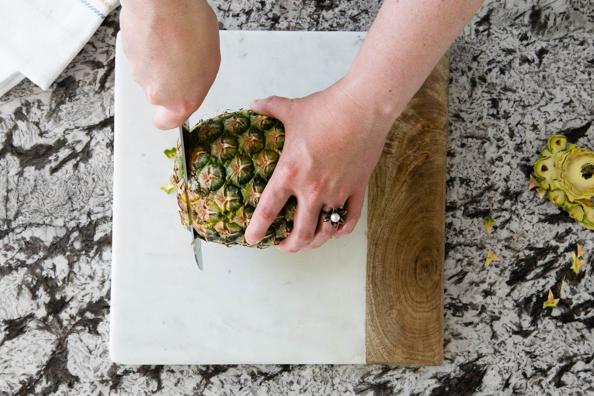 slicing off the top of a pineapple on a marble cutting board.
