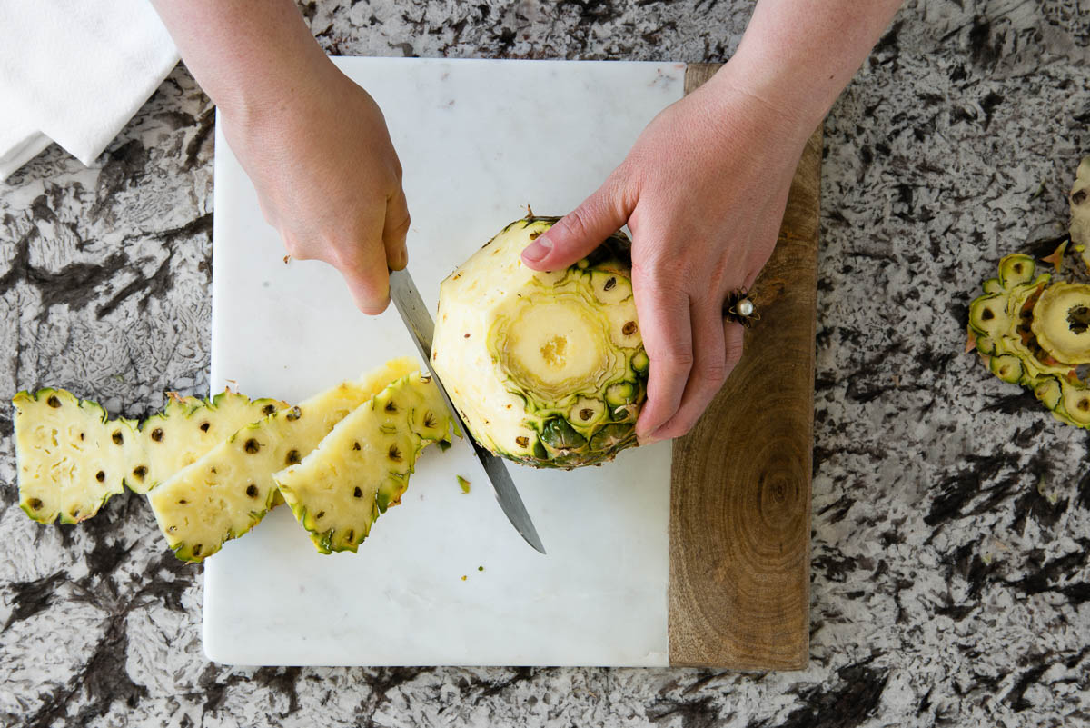 showing how to cut pineapple by removing the skin with a big vegetable cleaver on a marble cutting board.