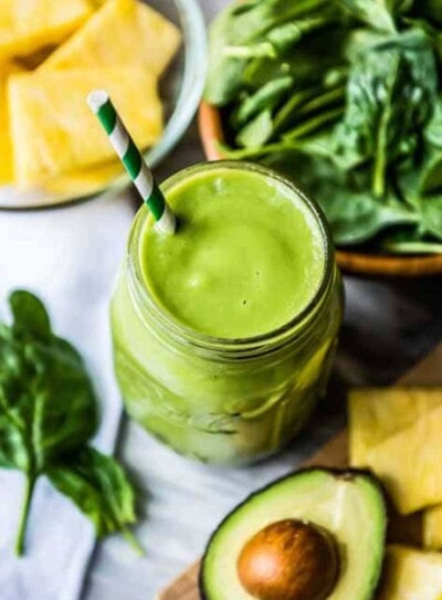 green clear skin smoothie in a mason jar with a paper straw, on a counter with avocado, pineapple and spinach.