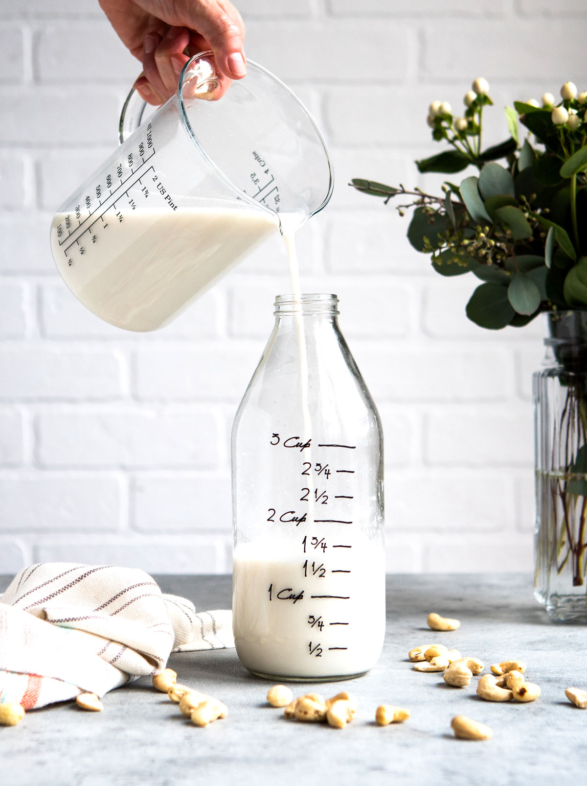 A hand pouring a homemade cashew milk recipe into a clear glass jar.