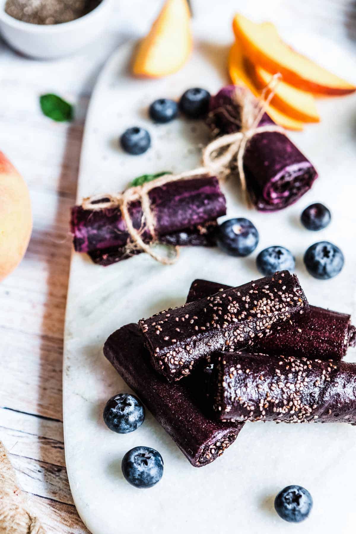 4 purple fruit roll ups with chia seeds on a white plate with fresh blueberries.