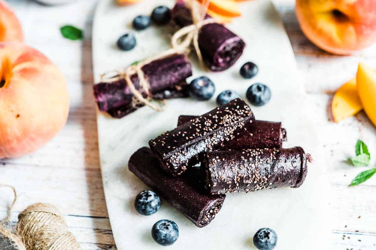 rolls of fruit leather on a marble serving tray surrounded by fresh blueberries and peaches.