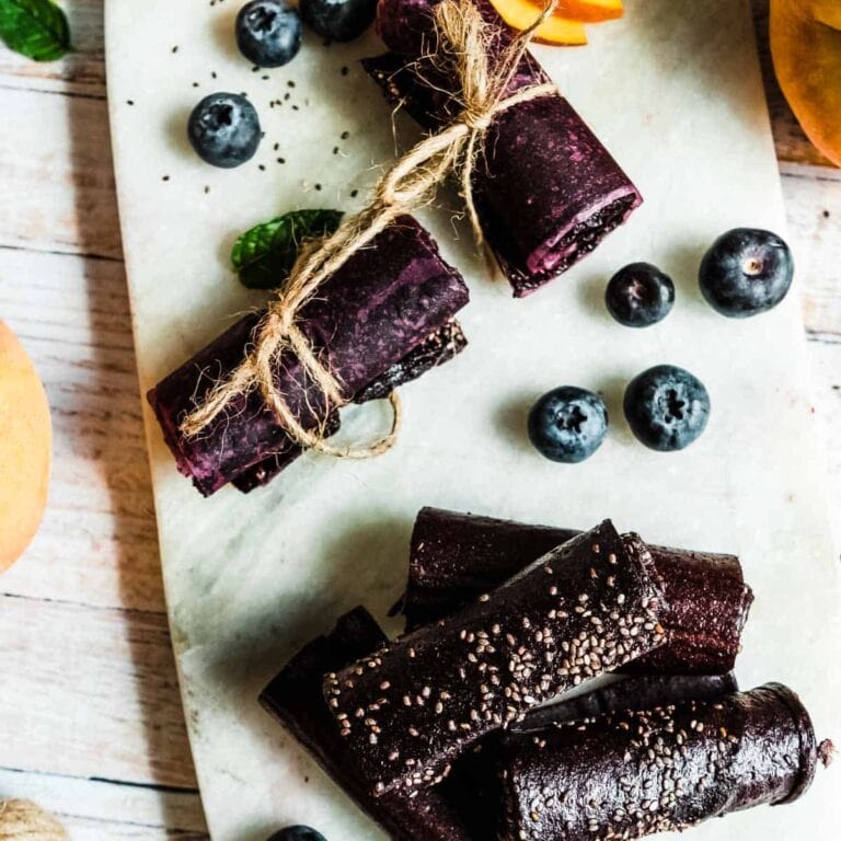 rolls of fruit leather on a marble serving tray surrounded by fresh blueberries and peaches.