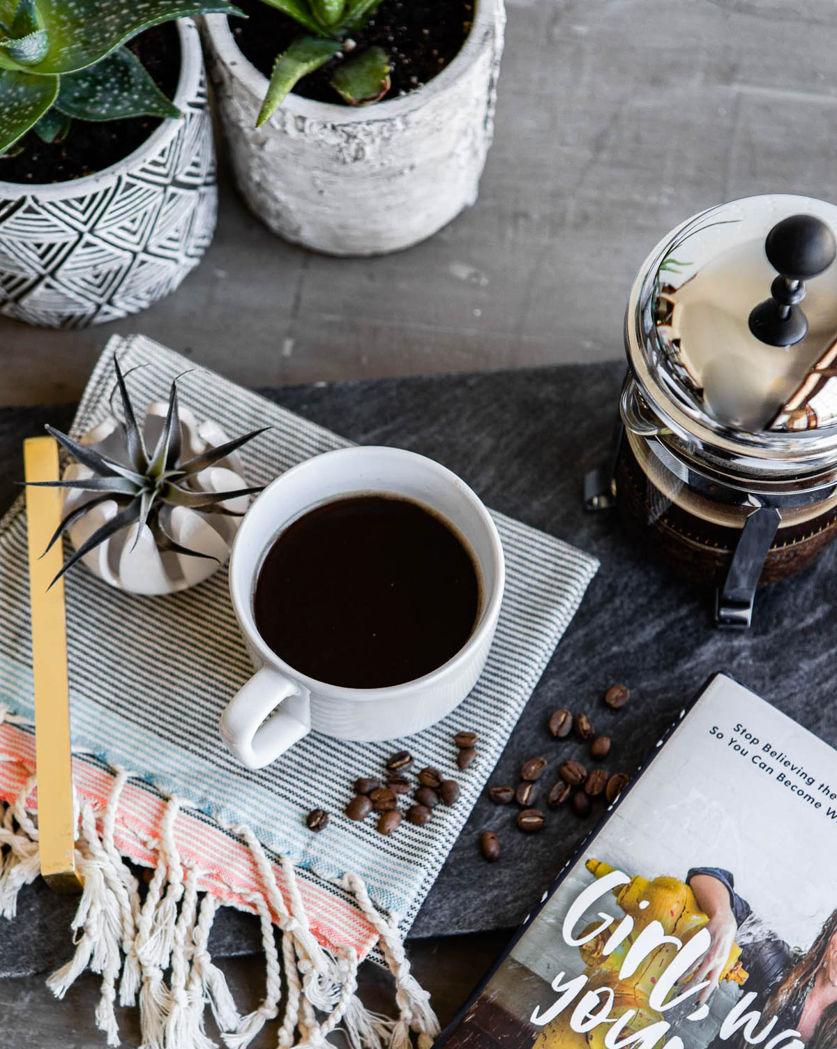 white cup of healthy coffee on a striped tea towel next to a french press and some whole coffee beans.