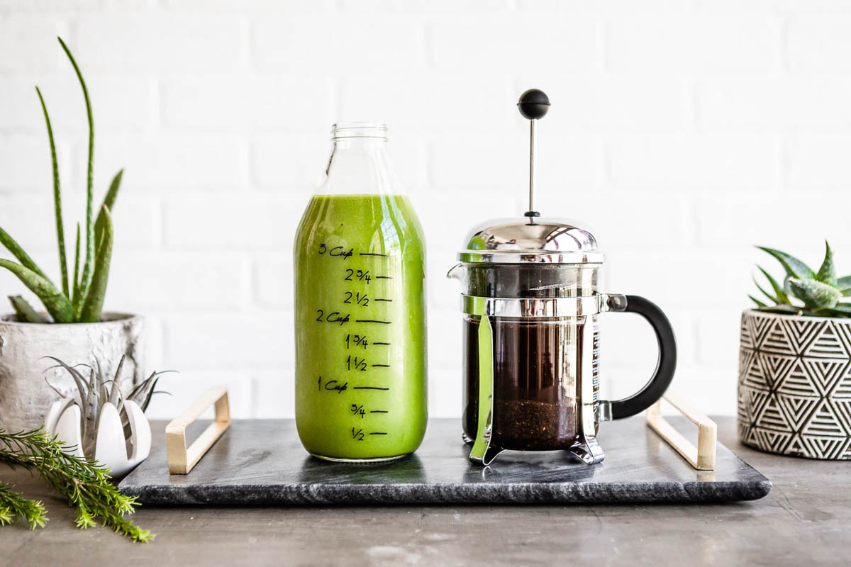 a large jar of green smoothie next to a french press both on a marble tray.