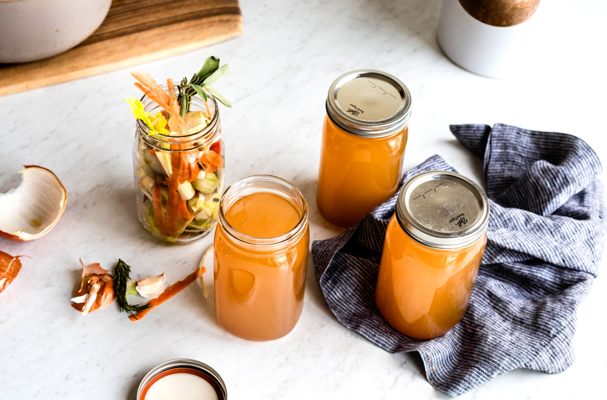 Mason jars of homemade vegetable stock with vegetable scraps on marble counter.