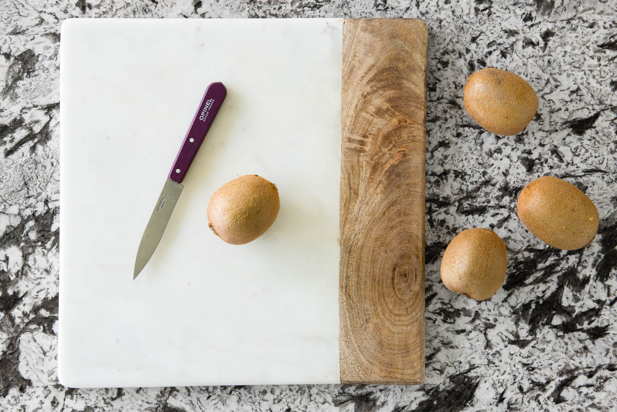 showing how to cut a kiwi using a marble and wood cutting board, a paring knife and 4 whole kiwis.