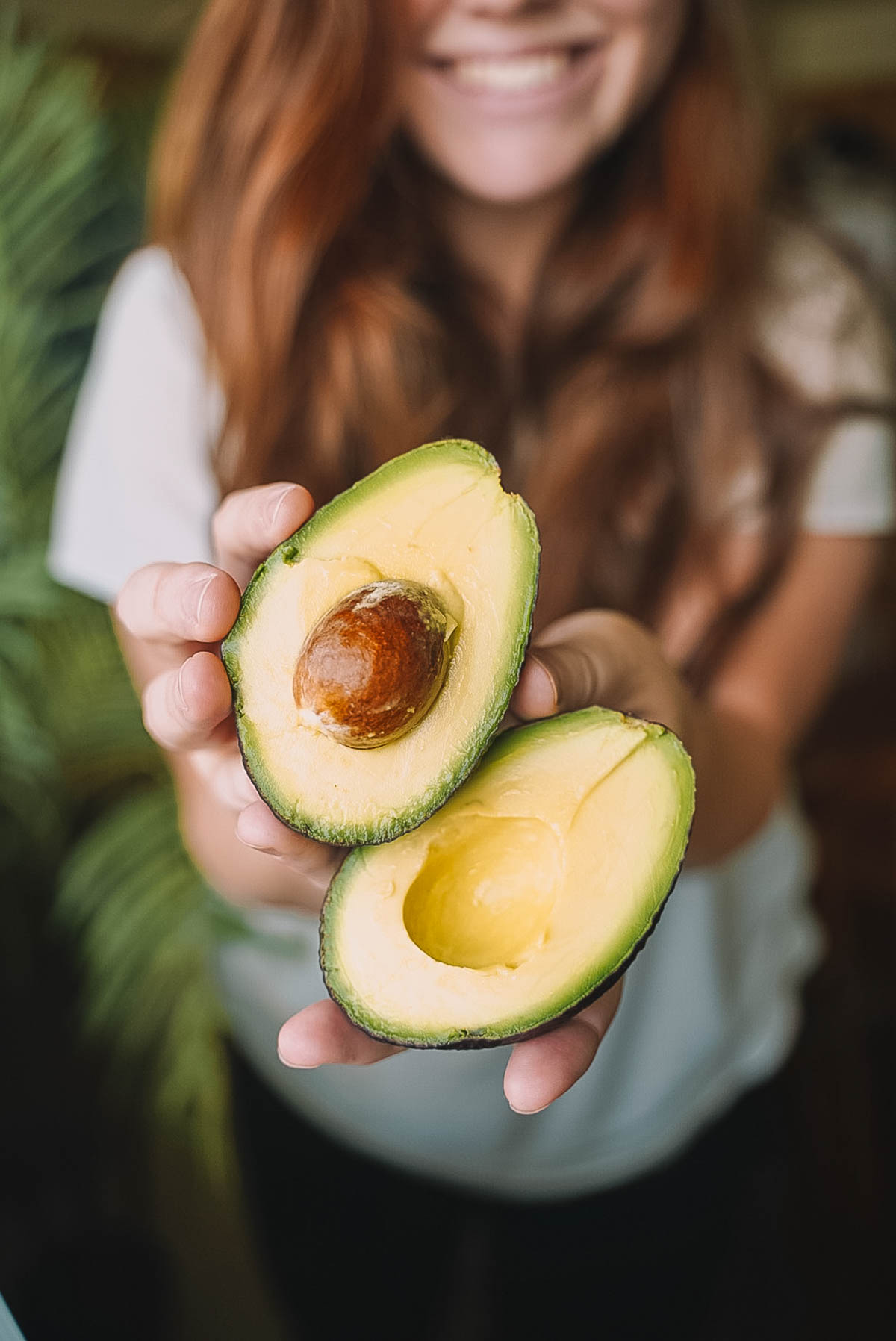 Ripe avocado sliced in half in 2 hands with lady smiling in the background.