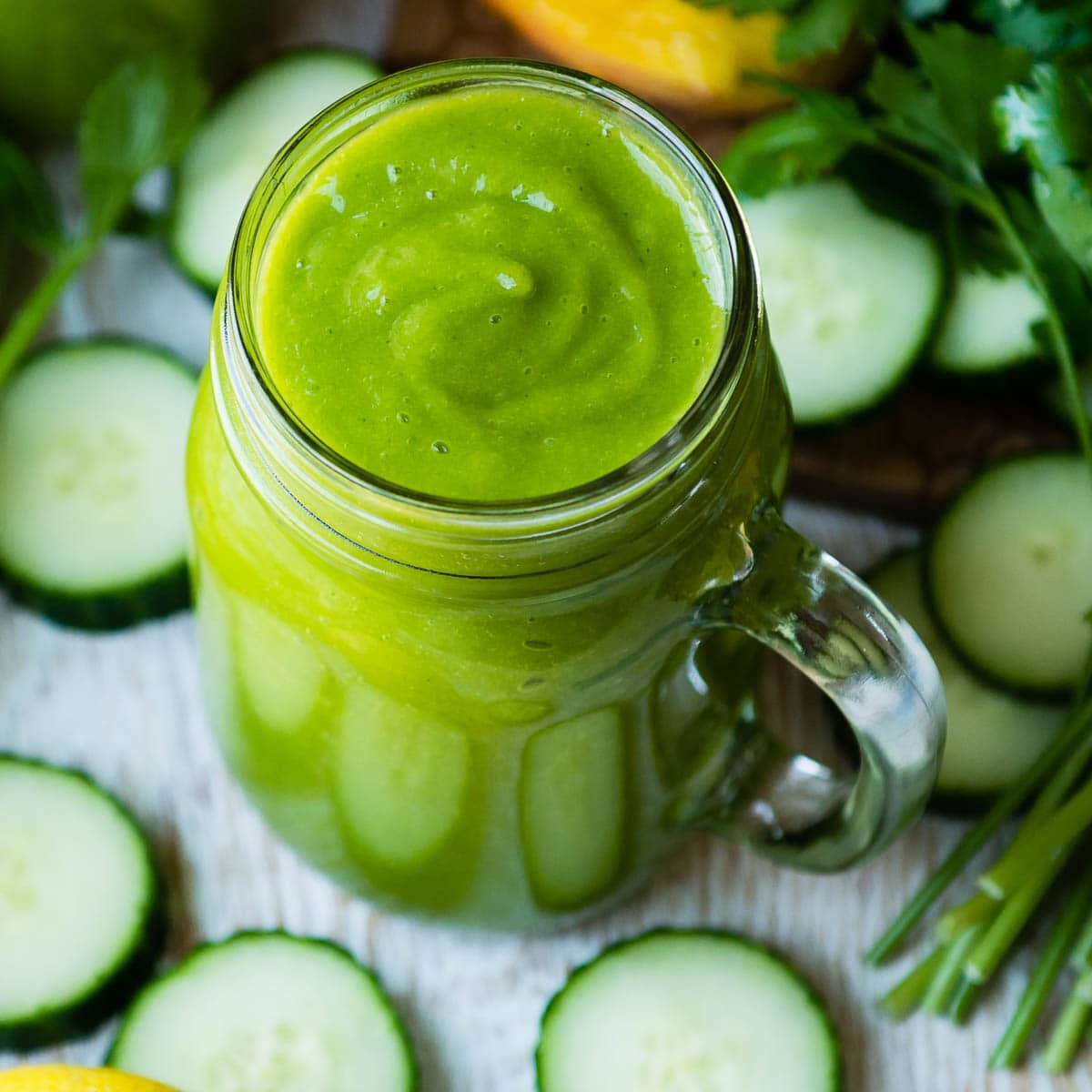 glass jar of green immune system booster smoothie surrounded by chopped cucumbers and parsley.
